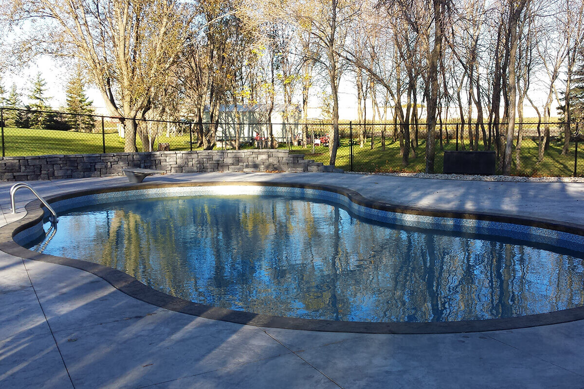 This eye level outdoor photograph captures a different perspective of the Waverley custom freeform pool with pavilion project, focusing on the calm water surface reflecting the surrounding nature. The curved freeform swimming pool takes up the central portion of the frame, detailed with a dark grey stone style border and a blue tile pattern along the inside perimeter. The clear water creates crisp reflections of the tall, bare trees lined up along the background property line. A massive textured concrete deck extends across the entire foreground, showing subtle shadows and clean seams. In the upper left background, a multi-tiered custom stone retaining wall built from dark grey blocks coordinates with a black chain-link safety fence that runs along the lawn. A stainless steel pool ladder with curved handrails is mounted on the far left edge of the pool coping, while a distant white building is visible through the tree line under a clear sky.