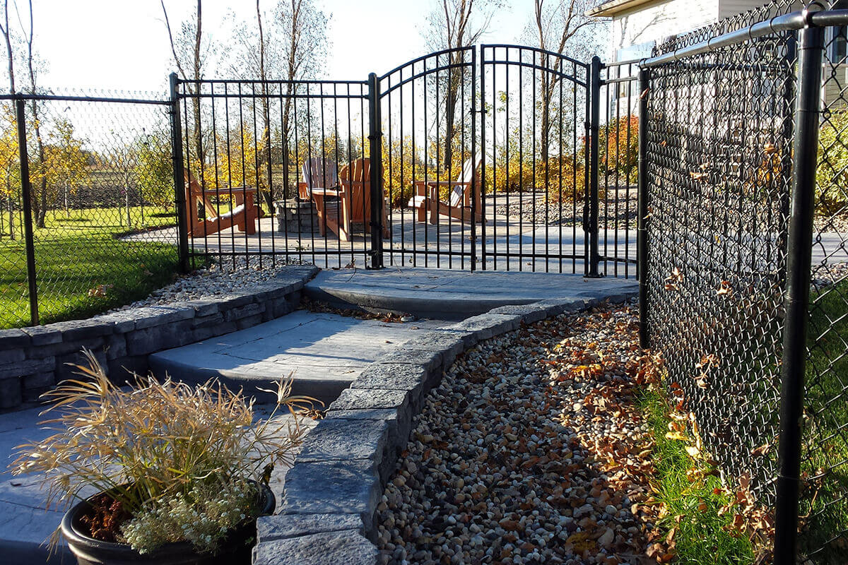 This close up outdoor photograph highlights the meticulous landscaping and safety fencing elements integrated into the Waverley custom freeform pool with pavilion property. In the centre background, a black arched metal safety gate stands closed, separating different zones of the backyard entertainment space. Beyond the gate, a stone patio features several wooden outdoor lounge chairs arranged around a central stone fire pit, bordered by a green lawn and autumn trees. In the foreground, a multi level walkway constructed from textured grey concrete steps is flanked by curved custom stone retaining walls. A dark plastic pot containing decorative green and tan grasses sits in the bottom left corner, while a wide drainage bed filled with tan river rock gravel and fallen brown leaves runs along the right side next to a black chain link fence.