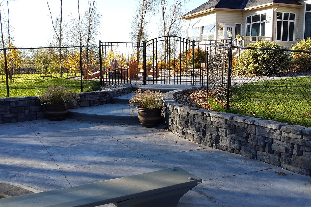 This elevated backyard photograph provides a broader view of the organized stone masonry and secure fencing layout that complements the Waverley custom freeform pool with pavilion installation. The foreground features a light grey textured concrete deck area with the tip of a grey diving board visible at the bottom centre. A multi-tiered custom stone retaining wall made of dark grey blocks curves upward on the right, holding two large potted decorative grasses on its ledges. Concrete steps lead up through a secure black arched metal safety gate that is built into a matching metal fence line. Beyond this secure perimeter, an upper stone patio reveals a group of wooden chairs surrounding a fire pit area, with a large beige two-story residential home featuring extensive white-trimmed windows standing prominently in the upper right background under a bright clear sky.