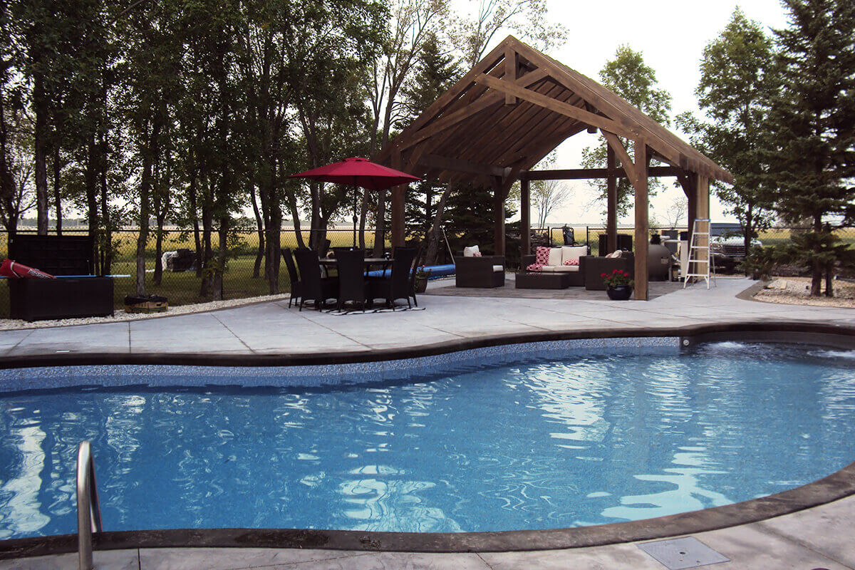 This mid range outdoor photograph offers a detailed view across the rippling water of the Waverley custom freeform pool with pavilion setup. The curved edge of the blue swimming pool dominates the foreground, framed by a smooth dark grey border and the light grey textured concrete deck. In the mid-ground on the left patio, a dark dining table with six matching chairs is shaded by a vibrant open red umbrella. To the right, the impressive heavy-timber wooden pavilion structure is showcased with its thick wooden posts and open rafter ceiling, shading an outdoor living area equipped with a cream-coloured sectional sofa and dark red throw pillows. A black utility storage box sits on the far left deck area against a backdrop of dense mature trees, while a white step ladder is visible near the back of the pavilion against an open country landscape background under a soft overcast sky.