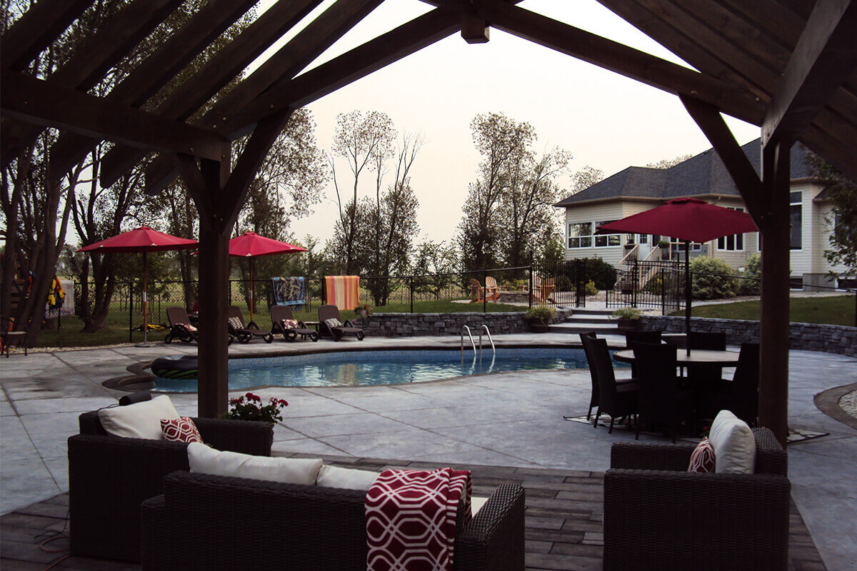 This interior perspective looks out from directly underneath the heavy-timber structure of the Waverley custom freeform pool with pavilion project, framing the entire outdoor living area. In the immediate foreground, dark brown wicker outdoor sofas with white cushions and red geometric patterned blankets fill the lower corners. Looking out past the thick wooden columns of the pavilion, the large textured concrete deck opens up to reveal the blue freeform swimming pool in the center of the yard, equipped with a stainless steel pool ladder. Across the pool deck, a patio dining table with a red umbrella sits to the right, while a row of dark lounge chairs under another red umbrella aligns against the far left fence line. A curved dark grey stone retaining wall separates the pool deck from an upper lawn, leading towards a large beige residential house with a dark shingled roof visible in the upper right background.