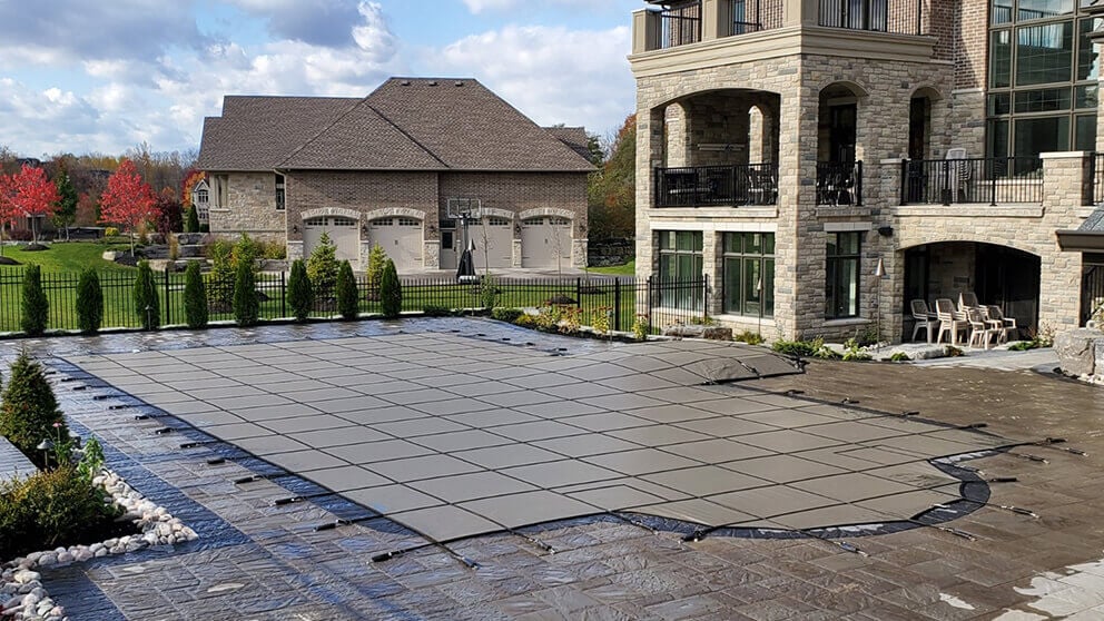 Installed grey PSP Pool Safety Cover fitted over a custom in-ground pool on a stone patio with deck tension straps.